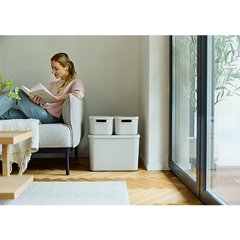 Femme assise sur un canapé lisant un livre, à côté de boîtes de rangement grises.
