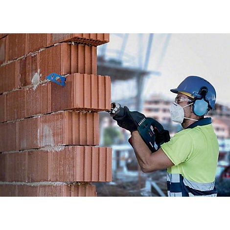Ein Bauarbeiter mit Schutzhelm und Maske sägt Ziegelsteine. Der Hintergrund zeigt eine Baustelle mit unscharfen Konturen.