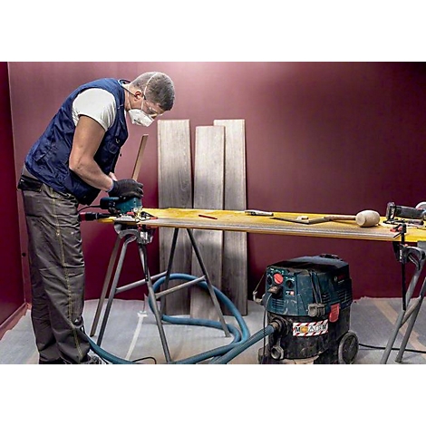 Handwerker schneidet Holz mit einer Kreissäge. Staubschutzmaske, Werkzeuge, hellgelbe Werkbank und grauer Boden. Roter Hintergrund.