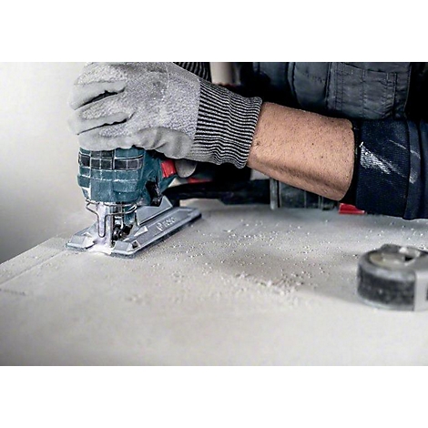 Person sägt mit einer Stichsäge ein Brett. Auf der Arbeitsfläche liegt Staub. Ein Maßband liegt am Rand.