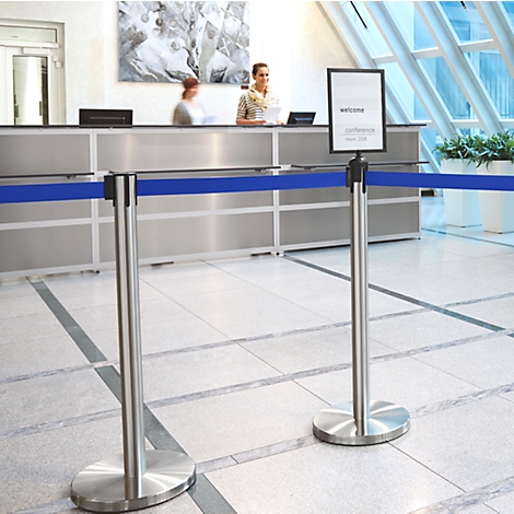 Reception desk with employees, barrier tapes and a welcome sign. The floor is light tiled. Windows and artwork in the background.
