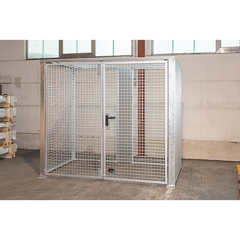 Une cage en treillis métallique argentée avec une porte et une serrure se trouve dans une pièce. Des fenêtres et un mur en béton sont visibles en arrière-plan.