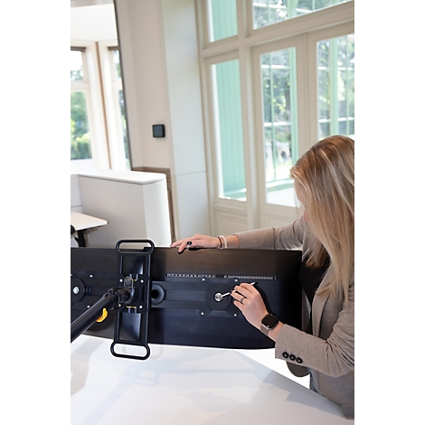 Une femme fixe un moniteur à un support avec une clé. Beaucoup de lumière du jour, de grandes fenêtres en arrière-plan.