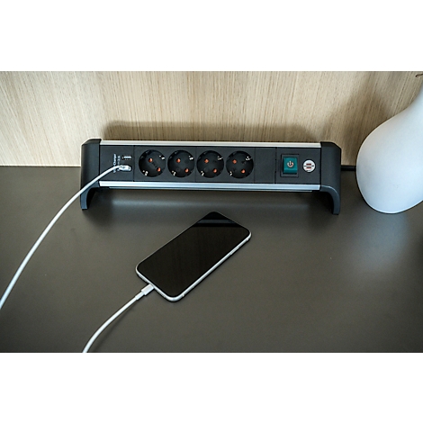 Multiprise noire avec 4 prises, à côté un smartphone avec câble de chargement. Un vase en arrière-plan.