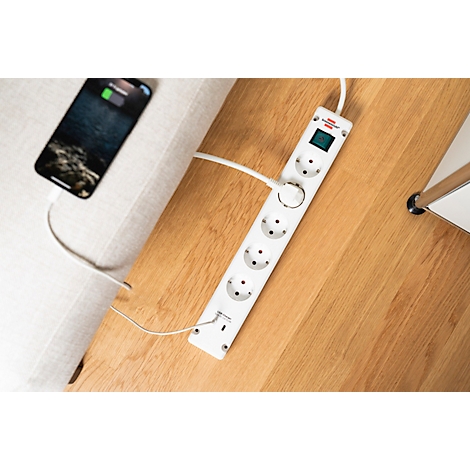 Een mobiele telefoon is via een wit verlengsnoer aangesloten op een witte stekkerdoos die op een houten tafel ligt.