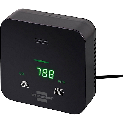 Een zwart apparaat met een groen display op een tafel. Op de achtergrond zit een persoon achter een laptop. Het display toont "751".