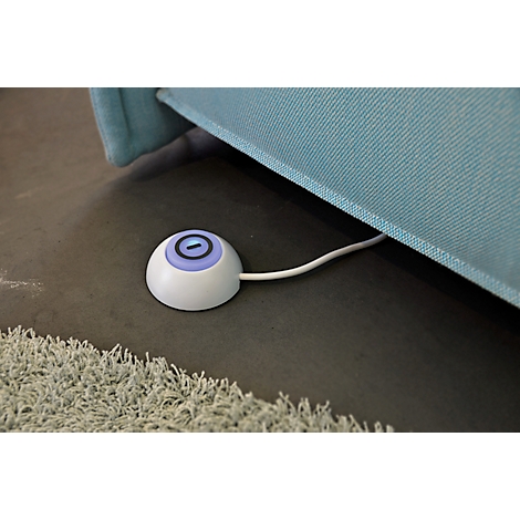 Appareil rond blanc au sol, avec un bouton bleu et un câble. Un canapé bleu est visible en arrière-plan.