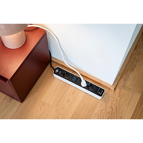 Une multiprise blanche avec écran est posée sur un sol en bois. Câbles d'alimentation et une lampe sont connectés.