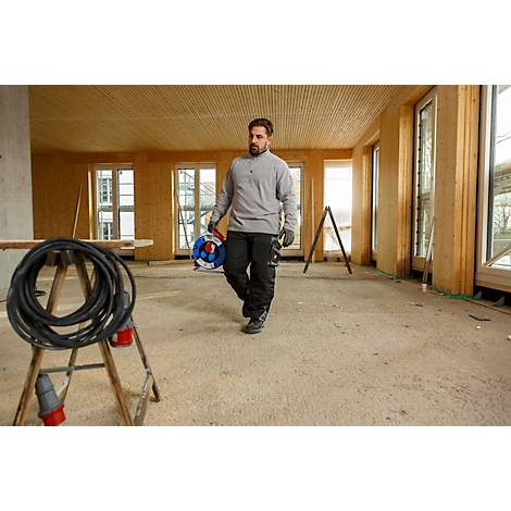 Ein Mann geht durch einen Raum mit Holzverkleidung, Kabeltrommel in der Hand. Im Vordergrund ein Kabel, links ein Tisch.