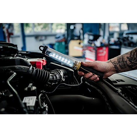 Une main tatouée tient une lampe LED brillante dans un compartiment moteur.