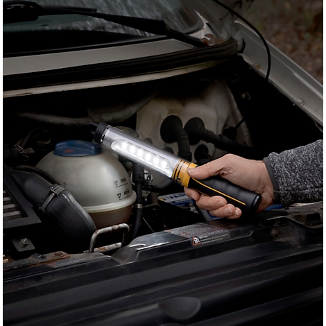 Une personne tient une lampe LED dans le compartiment moteur d'une voiture. La lampe est allumée. Le compartiment moteur est ouvert.