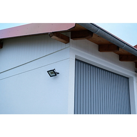 Lampe d'extérieur brillamment éclairée sur un mur gris au-dessus d'une porte. La lampe est carrée et émet une lumière vive.