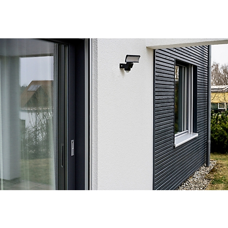 Buiten aan een muur van een huis staat een zwarte bewegingsmelder en een buitenlamp. Rechts een raam en grijze houten panelen.