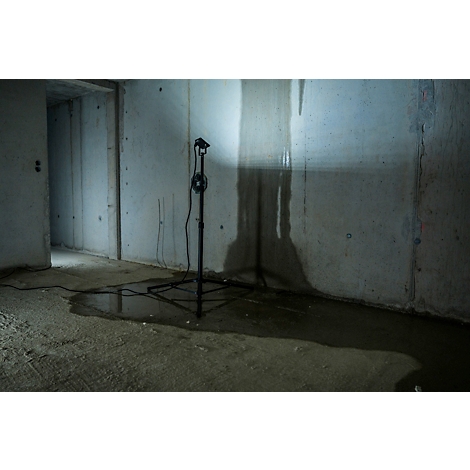 Betonnen kamer met plas op de vloer. Een zwart statief met lamp staat ervoor.