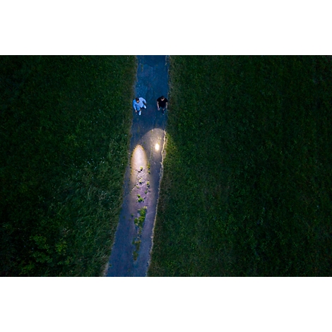 Zwei Personen gehen auf einem beleuchteten Weg durch eine grüne Wiese. Vogelperspektive.