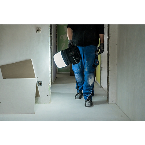 Persoon draagt lamp door gang in ruwbouw, blauwe werkkleding.