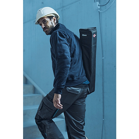 Un ouvrier du bâtiment en vêtements de travail et un casque porte un sac noir sur son épaule en montant un escalier.