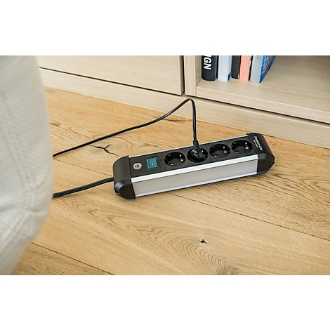 Une multiprise noire est posée sur un plancher en parquet. Une prise est branchée.