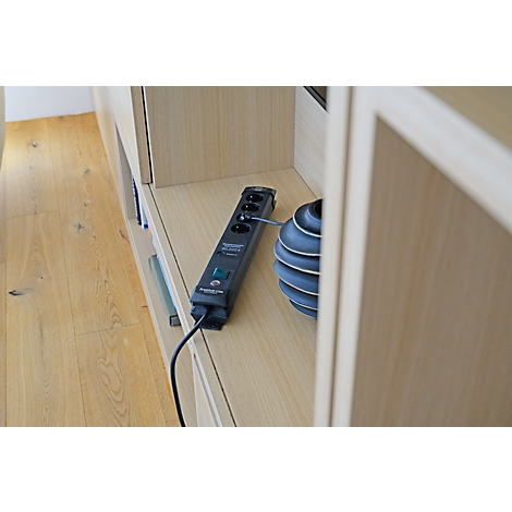 Een zwarte stekkerdoos met 4 stopcontacten, een schakelaar en een stroomkabel. Hij ligt op een lichte houten plank naast een decoratief, blauw voorwerp.