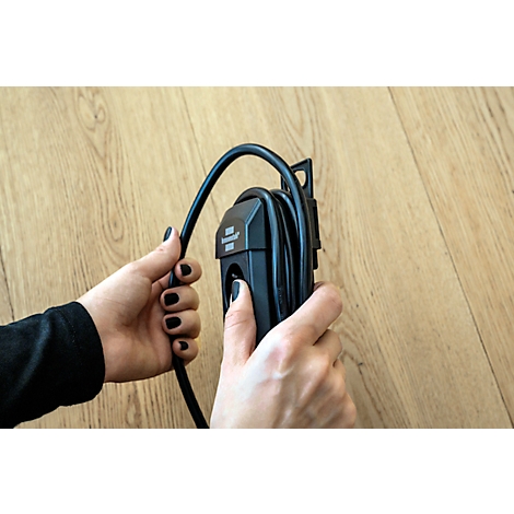 Mains tenant un câble de chargement noir pour une voiture électrique. Le fond est un plancher en bois.
