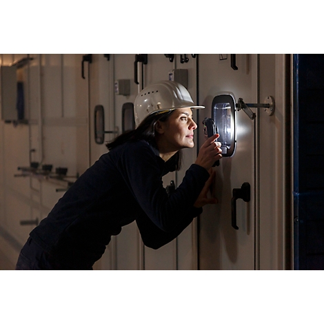 Een vrouw met een helm onderzoekt een schakelpaneel met een lamp. Ze lijkt geconcentreerd.