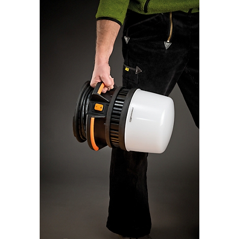 Een persoon draagt een mobiele bouwplaatslamp, bestaande uit een witte lampenkap en een zwarte behuizing. De handgreep is oranje.