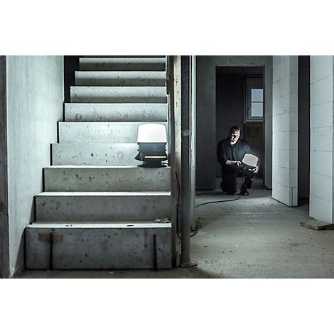 Un homme s'agenouille sur un chantier et tient une lampe de travail brillante. Escalier et lampe à droite, gros œuvre à gauche.
