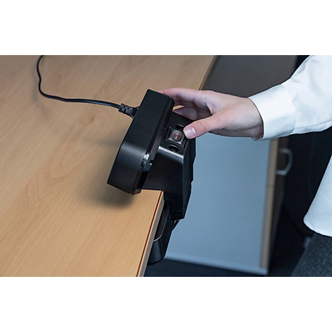 Een persoon drukt op een rode knop op een zwart apparaat dat aan een bureau is bevestigd.