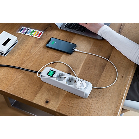 Nahaufnahme: Eine Hand öffnet eine Klappe einer schwarzen Steckdosenleiste mit USB-Anschlüssen. Der Hintergrund ist Holzoptik.