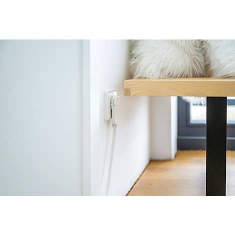 Close-up van een wit stopcontact aan een muur. Een witte kabel hangt naar beneden. Op de achtergrond: houten tafel met kussens.