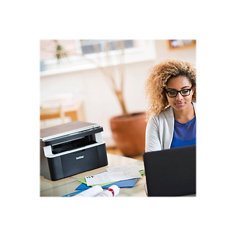 Eine Frau mit Brille sitzt vor einem Laptop. Daneben ein Drucker, Papiere und eine Topfpflanze.