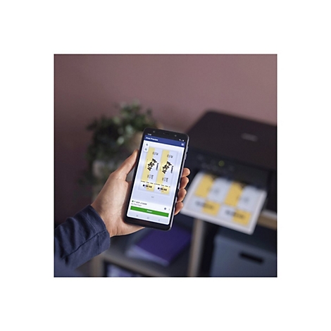 Eine Hand hält ein Smartphone mit Druckvorschau. Ein Drucker ist im Hintergrund zu sehen. Auf dem Smartphone-Bildschirm werden zwei gelbe Etiketten angezeigt.