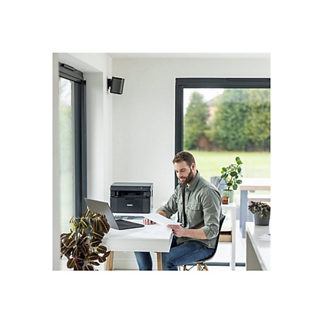 Ein Mann sitzt an einem Schreibtisch, liest Papier. Auf dem Tisch sind Laptop und Drucker.
