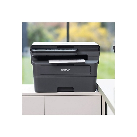 Ein schwarzer Brother-Multifunktionsdrucker steht auf einem weißen Tisch. Das Papier kommt aus dem Drucker. Im Hintergrund ist verschwommenes Grün zu sehen.