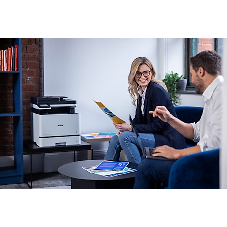 Eine lächelnde Frau und ein Mann arbeiten in einem modernen Büro zusammen, neben ihnen steht ein Brother-Drucker.