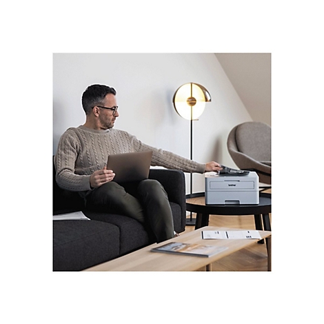 Ein Mann sitzt auf einem Sofa und bedient einen Drucker. Er hält einen Laptop und legt Papiere in den Drucker ein.