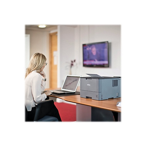 Frau sitzt am Schreibtisch, vor ihr Laptop und Drucker. Im Hintergrund TV.