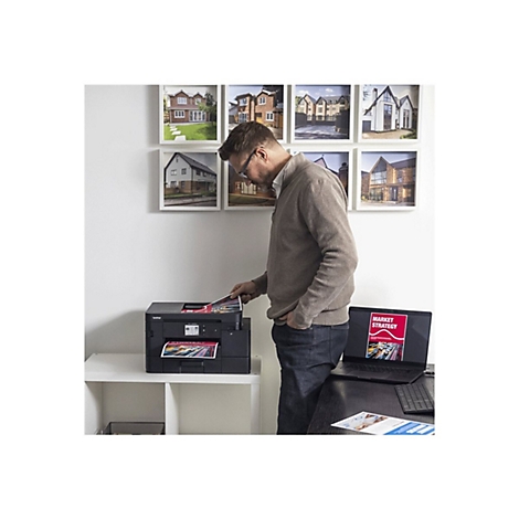 Ein Mann legt ein gedrucktes Dokument in einen Drucker. Im Hintergrund hängen gerahmte Hausfotos. Auf dem Tisch steht ein Laptop mit der Aufschrift "Market Strategy".