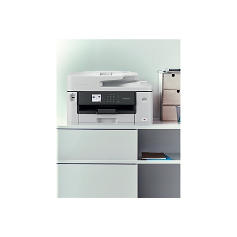 Ein weißer Multifunktionsdrucker steht auf einem Regal. Auf dem Display des Druckers werden Informationen angezeigt. Rechts davon ist ein Bücherstapel und Schubladen.