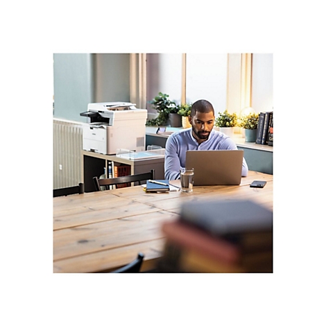 Ein Mann sitzt an einem Tisch und arbeitet an einem Laptop. Im Hintergrund befindet sich ein Drucker und Bücher.