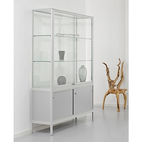 Een moderne, elegante vitrinekast van glas en metaal staat in een minimalistische ruimte. Beneath zijn er twee grijze lades en boven zijn er glazen planken met verschillende vazen erop. Rechts van de kast staat een artistieke houten stoel.