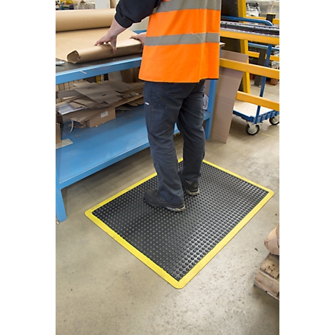 Person stands on a black mat with a yellow border in a factory. They are wearing a vest.