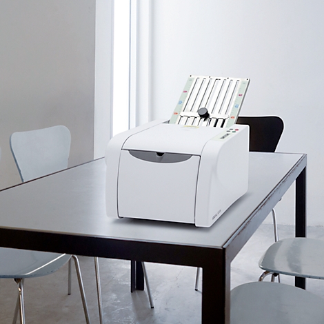Weißer Drucker mit offenem Fach steht auf einem Tisch. Im Hintergrund Stühle, Fenster.