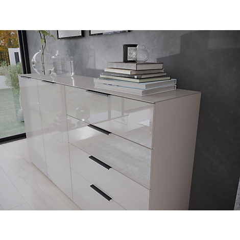 Un buffet crème brillant, avec des livres et de la décoration dessus. Poignées noires sur les tiroirs et les portes. En arrière-plan, des fenêtres avec vue sur l'extérieur.