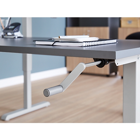 Bureau gris avec manivelle pour le réglage en hauteur. Des blocs-notes et des stylos sont sur le plateau.