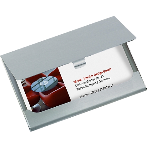 Silver business card holder displaying a business card with the Merlo. Interior Design GmbH logo. In the background, red armchairs are visible.