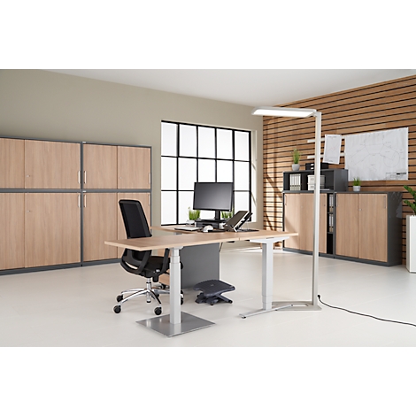 An office room with desk, chair, and cabinets. On the desk a computer, a lamp, and a footrest. Light wood and grey accents.