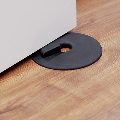 Un butoir de porte en caoutchouc noir devant une porte blanche sur un sol en bois. Le butoir de porte a une forme ronde avec un trou et une saillie.