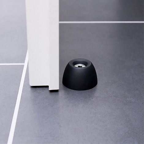 Un butoir de porte noir devant un cadre de porte blanc. Sur un sol gris. Le détail montre la forme de l'arrêt.