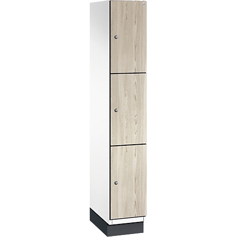 Smalle, driedeurs locker kast, houtlook, zilverkleurige knoppen. Witte zijwand, donkere sokkel.